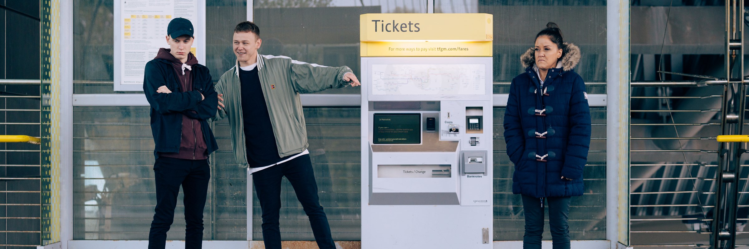 Two men point and laugh at a woman at a Metrolink tram stop.